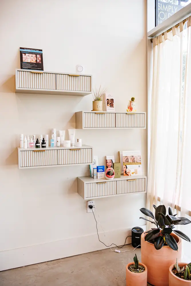 A minimalist room features wall shelves with skincare products, potted plants, and a sunlit window with sheer curtains next to a white wall.