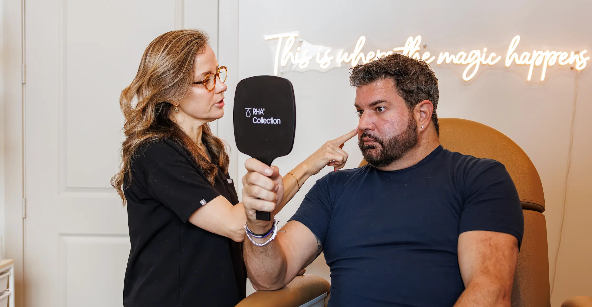 Two people are interacting in a room with neon text reading "This is where the magic happens." One holds a mirror.