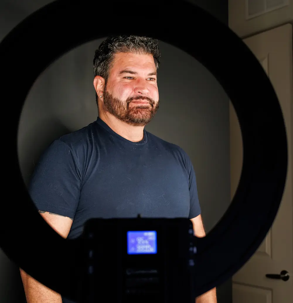 A person stands inside a photo studio, illuminated by a circular ring light, with a camera capturing the scene from the front.