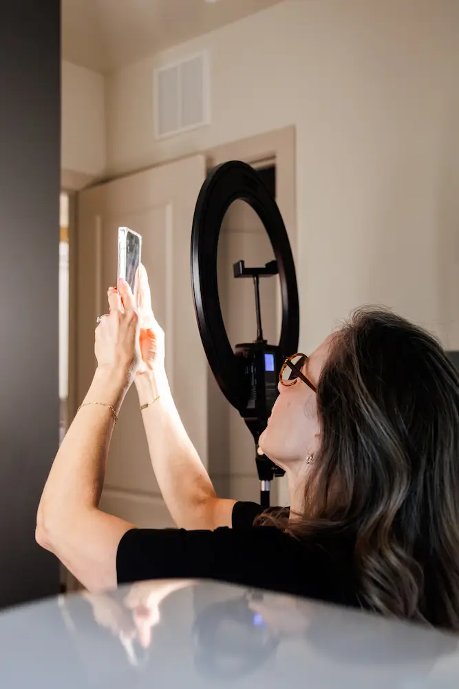 A person takes a photo with a smartphone, using a ring light for illumination, in a plain indoor setting.