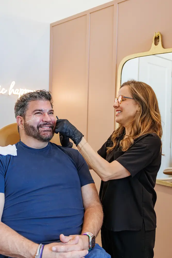 A person in gloves examines another's face, possibly a cosmetic consultation, in a warmly lit room with a mirror and soft-colored walls.