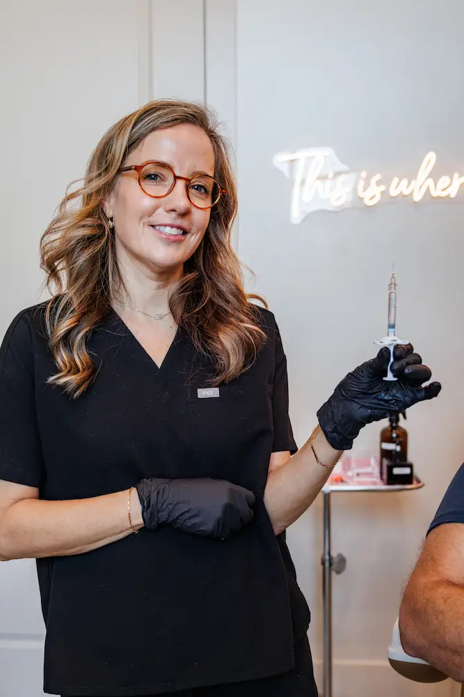 A person wearing glasses and black gloves holds a syringe, standing in front of a blurred neon sign and medical supplies.