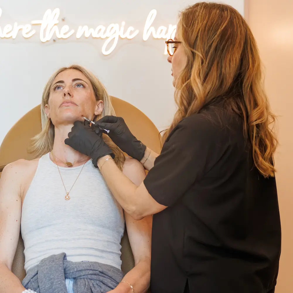 A person receives a cosmetic procedure from a professional in a modern clinic. A neon sign in the background reads, “where the magic happens.”