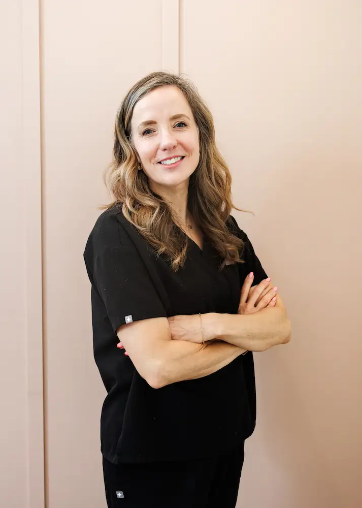 A person with long hair in black attire stands confidently, arms crossed, against a plain, light-colored wall.