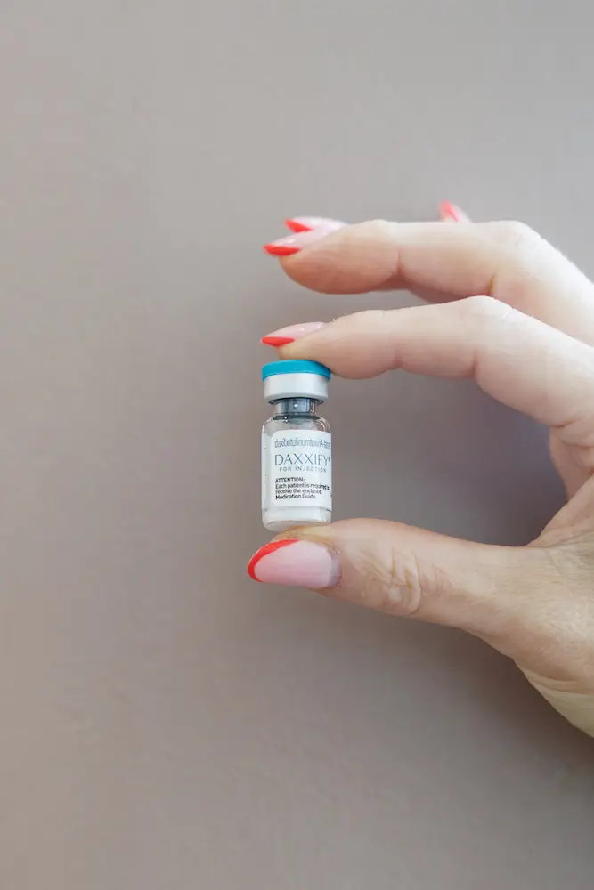 A person's hand with red nails holds a small vial labeled "Daxxify" against a neutral background, focusing on beauty or medical use.
