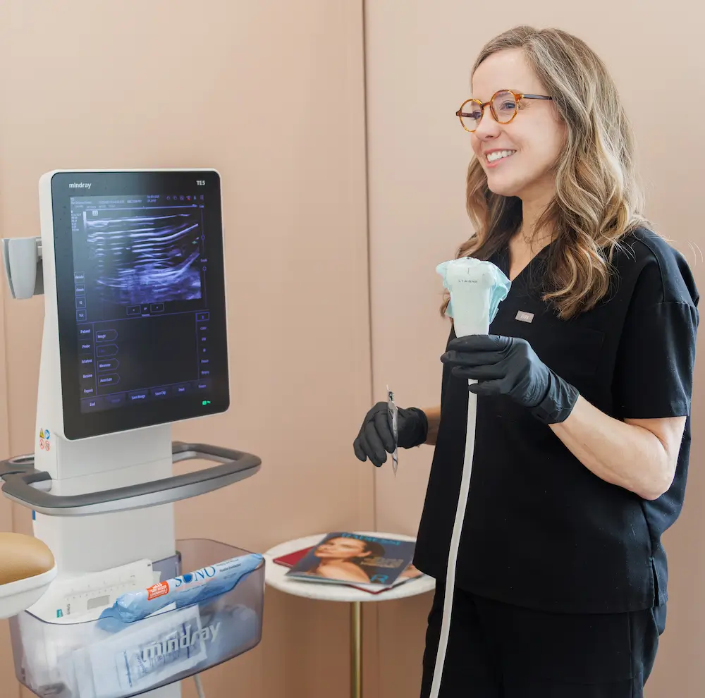 A person wearing gloves holds a device next to an ultrasound machine. A table with magazines and supplies is nearby.