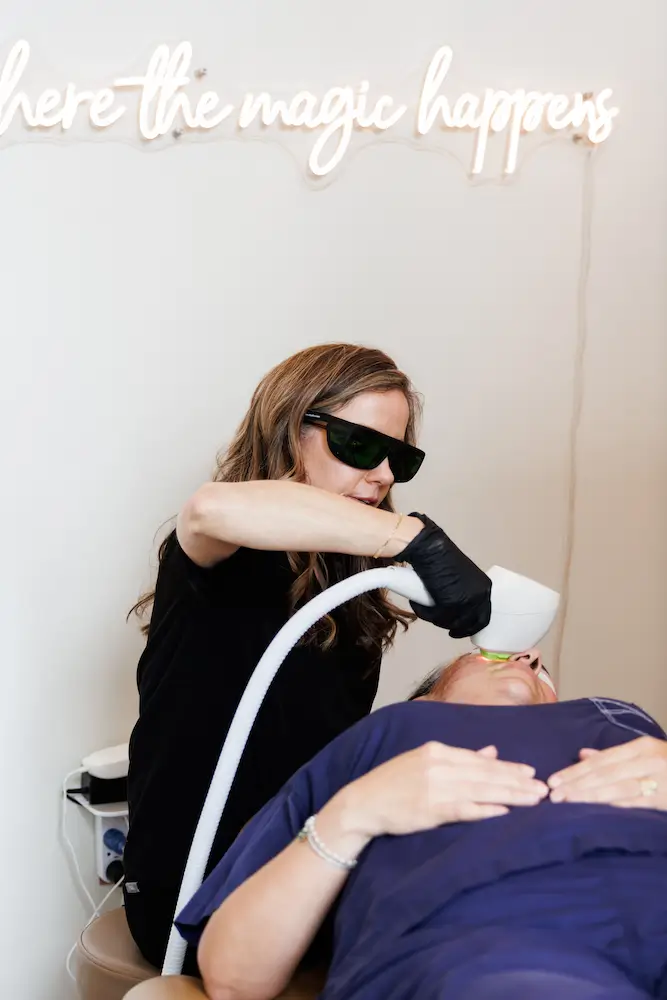 A person wearing protective glasses uses laser equipment on another person, beneath a neon sign reading "where the magic happens" in a medical setting.
