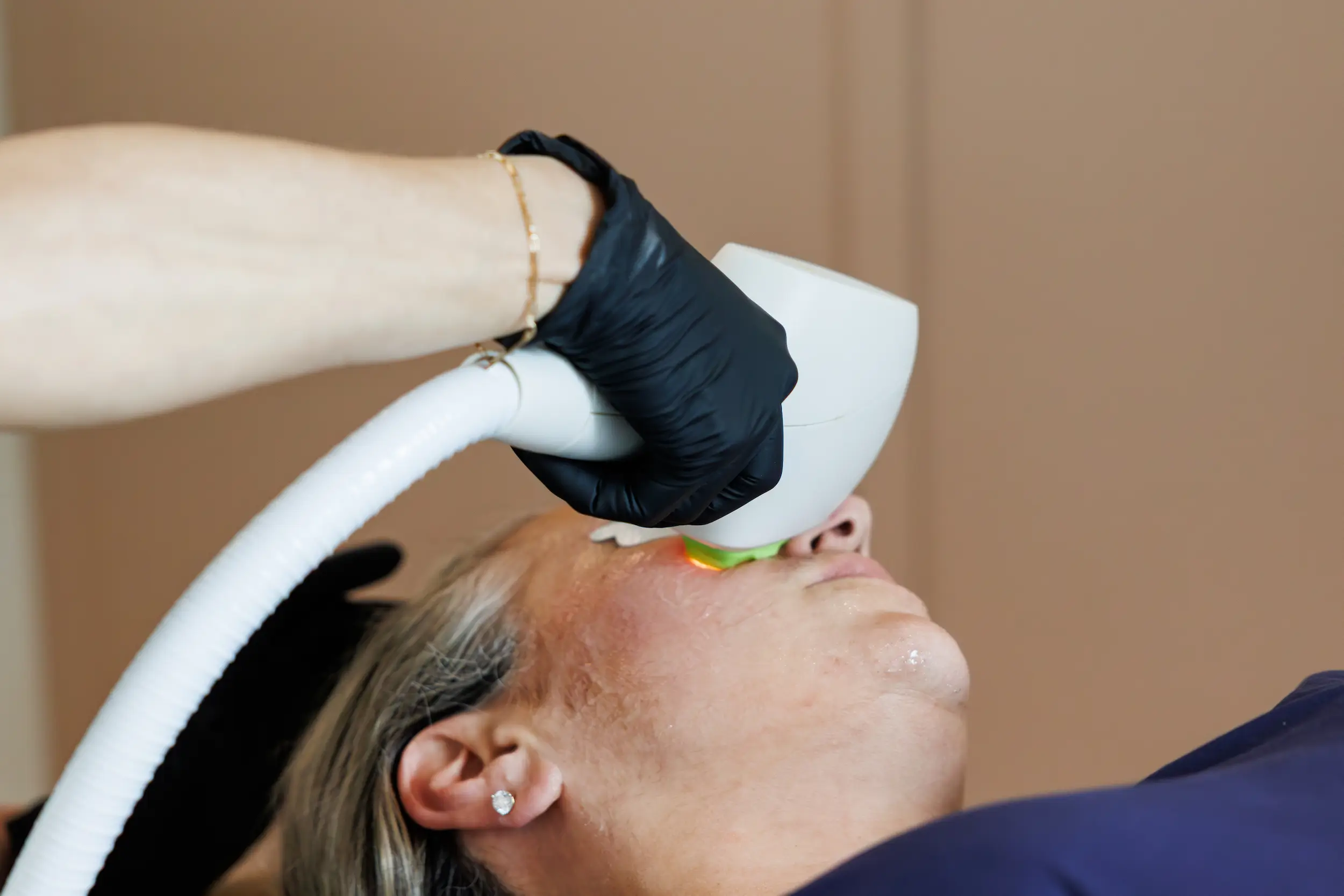 A person receives a facial treatment using a handheld device operated by another person wearing black gloves, focusing on the eye area.
