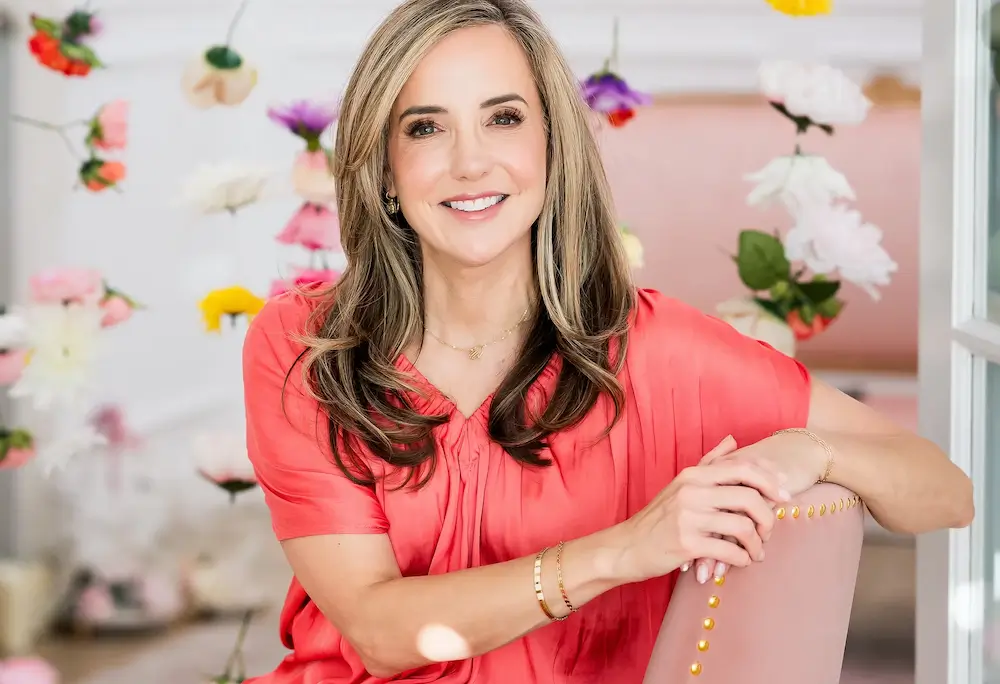 A person in a red blouse is smiling indoors, surrounded by hanging flowers, leaning on a pink chair with decorative pins.