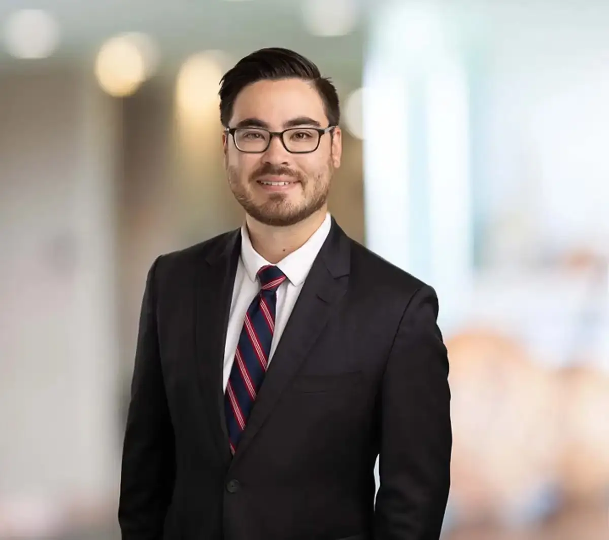 A person in a suit and tie stands indoors with a blurred background, smiling gently, glasses reflecting light. No landmarks visible.