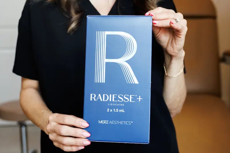 A person holds a blue Radiesse+ box with lidocaine, featuring the brand logo and product details, inside a well-lit room.