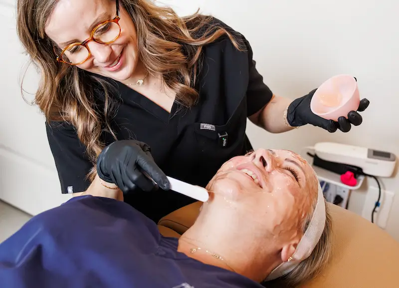 A person smiles while applying a facial treatment to another person lying down in a spa or clinic setting.