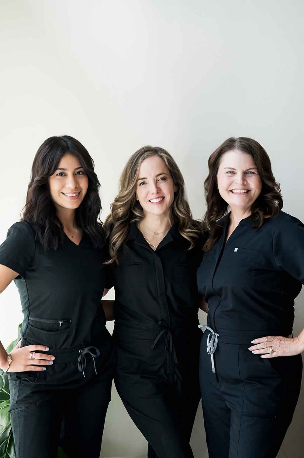 Three people in black attire stand together, smiling, against a plain white background. The setting is bright and minimalistic.