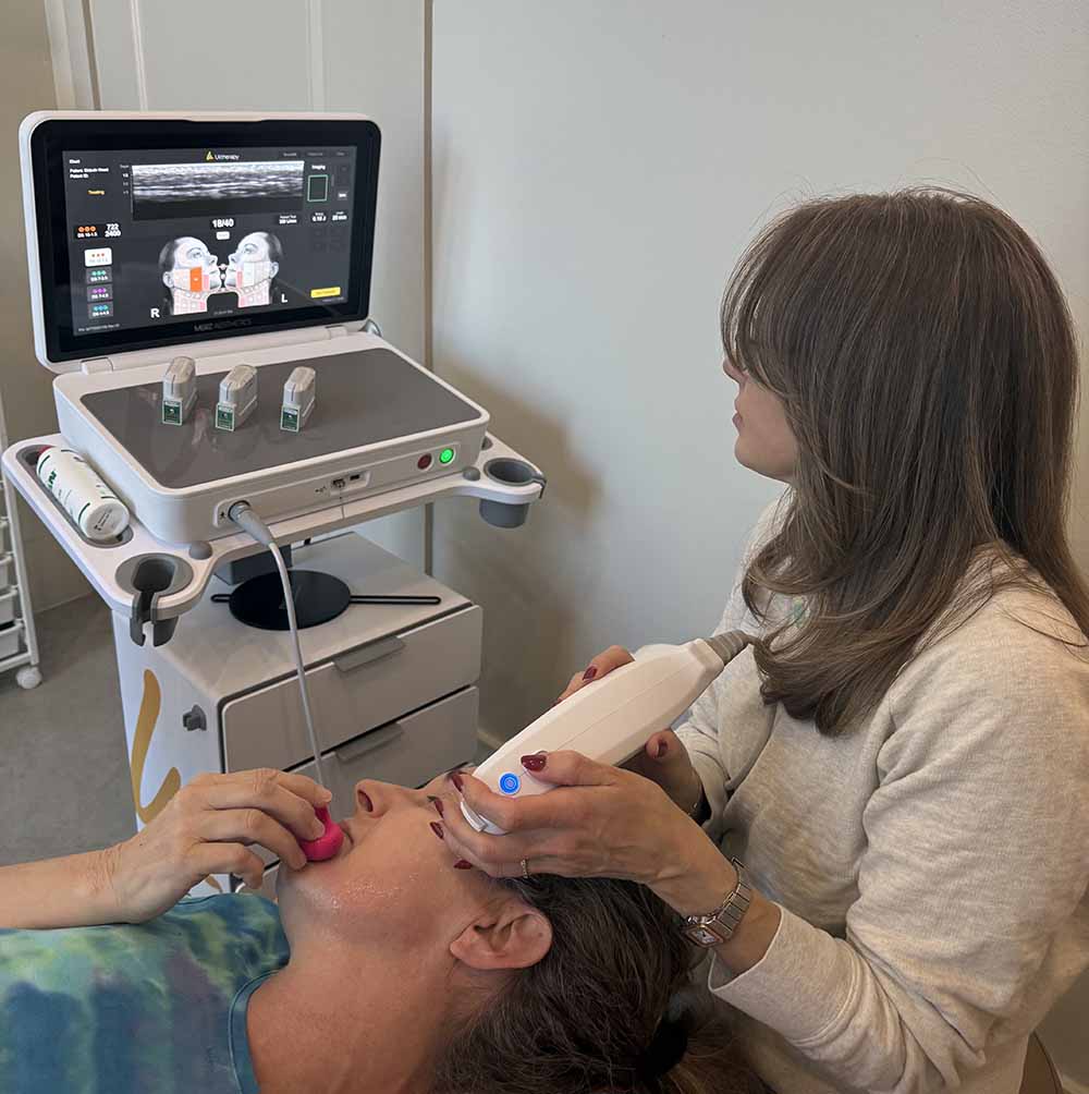 A person uses a medical device on another person’s face, with a monitor displaying facial images, in a clinical setting.